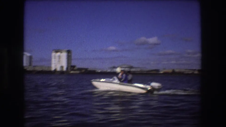 1975 Boston home-movie frame showing a white outboard motorboat with visible passengers moving across harbor water, with industrial shoreline structures in the distance.