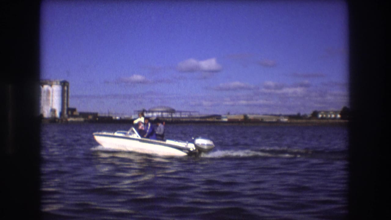 Small white motorboat moves across open water with shoreline structures behind it.