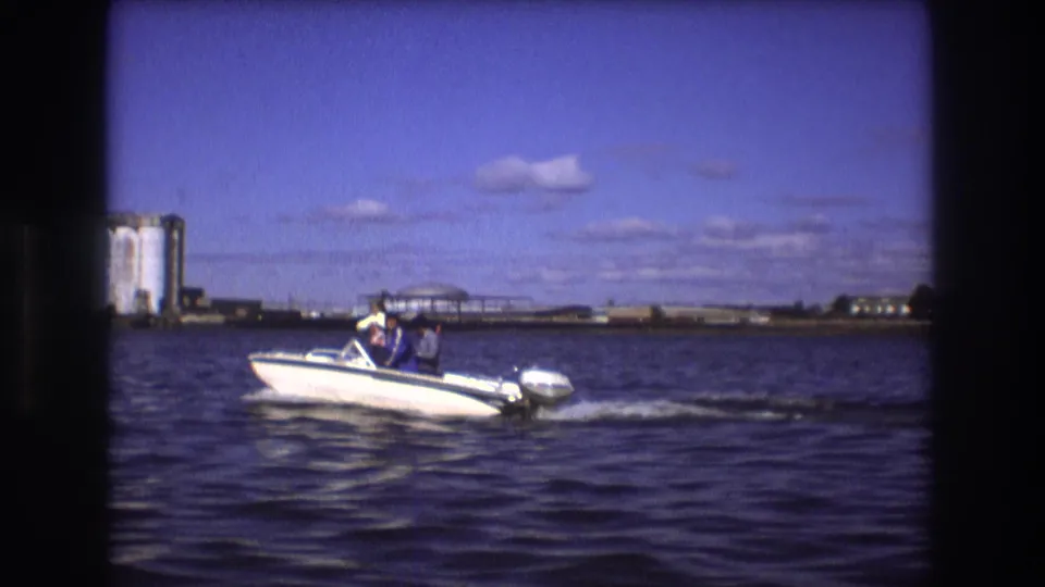 1975 Boston waterfront frame with a small white motorboat and passengers on rippled harbor water, set against distant industrial buildings and low shoreline structures.