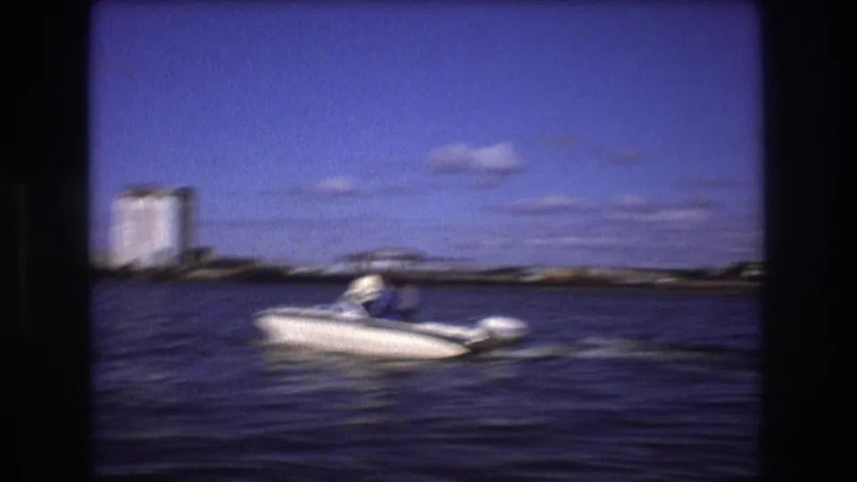 1975 Boston home-movie frame with motion blur as a white outboard motorboat passes on dark harbor water, with distant waterfront buildings under a blue sky.