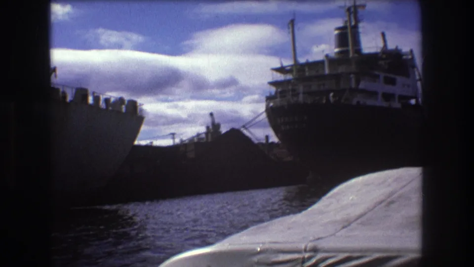 1975 Boston harbor frame showing a large dark-hulled working vessel beside the water, with a white boat covering or bow area in the foreground and clouds overhead.