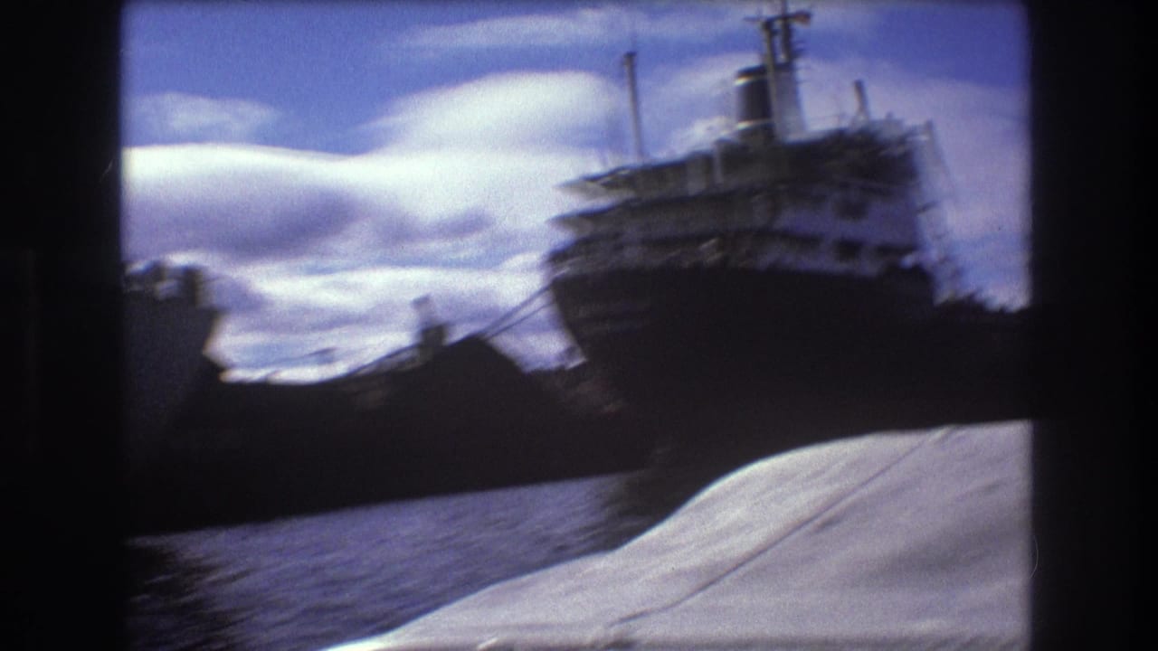 1975 Boston harbor home-movie frame showing a large dark-hulled cargo ship beyond water, filmed from a nearby boat with a pale foreground deck or canopy.
