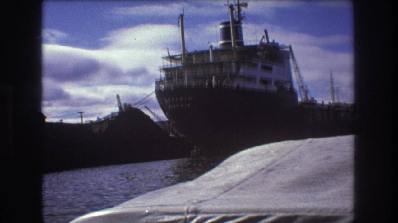 1975 Boston harbor frame with a docked cargo ship, white superstructure, dark hull lettering, water, and the filming boat's foreground deck.