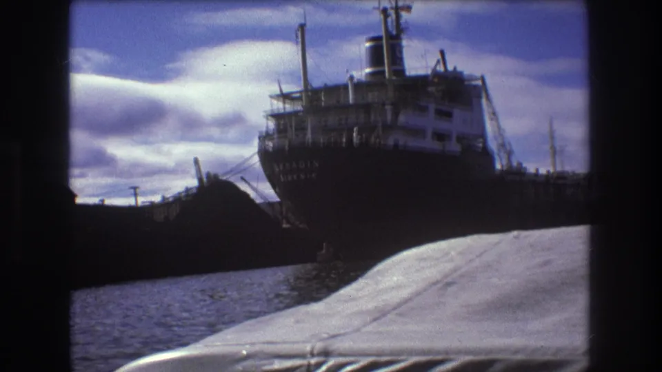 1975 Boston harbor frame with a docked cargo ship, white superstructure, dark hull lettering, water, and the filming boat's foreground deck.
