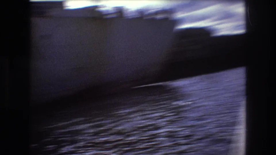 Motion-blurred 1975 Boston harbor frame showing the dark side of a large ship, rippled water, and dockside shapes during handheld filming from a boat.