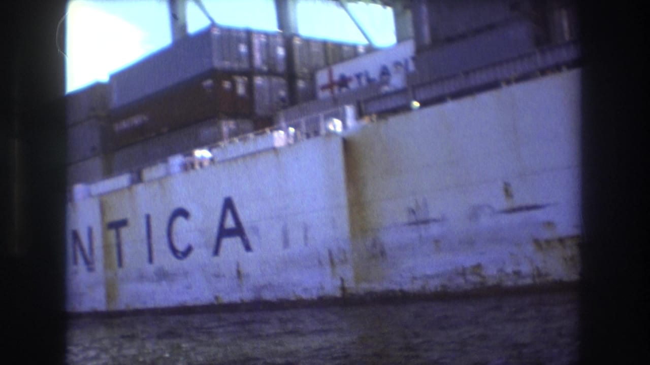 1975 Boston harbor home-movie frame showing the rust-stained white hull of a docked freight ship with partial ATLANTICA lettering and port structures above.
