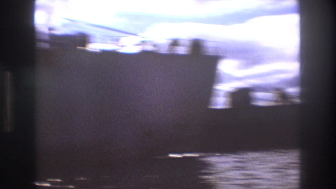 Color-faded 8mm frame of a large freighter-like vessel in Boston harbor, with water in the foreground and cloudy sky above.