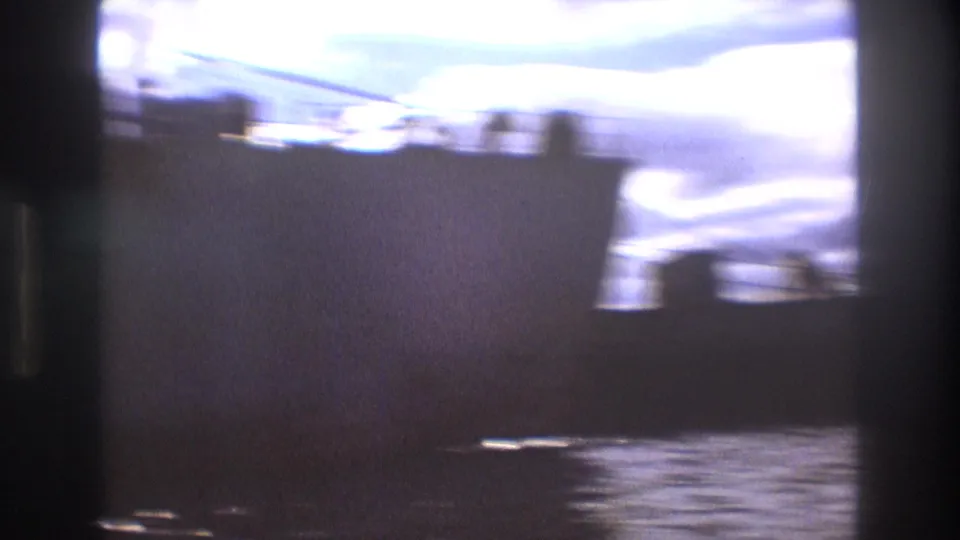 Color-faded 8mm frame of a large freighter-like vessel in Boston harbor, with water in the foreground and cloudy sky above.