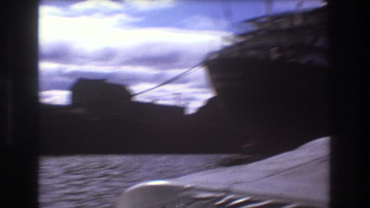 Handheld 8mm view across Boston harbor showing a dark vessel shape, rippling water, cloudy sky, and a pale bow or deck edge in the foreground.