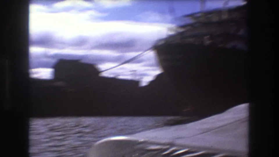Handheld 8mm view across Boston harbor showing a dark vessel shape, rippling water, cloudy sky, and a pale bow or deck edge in the foreground.