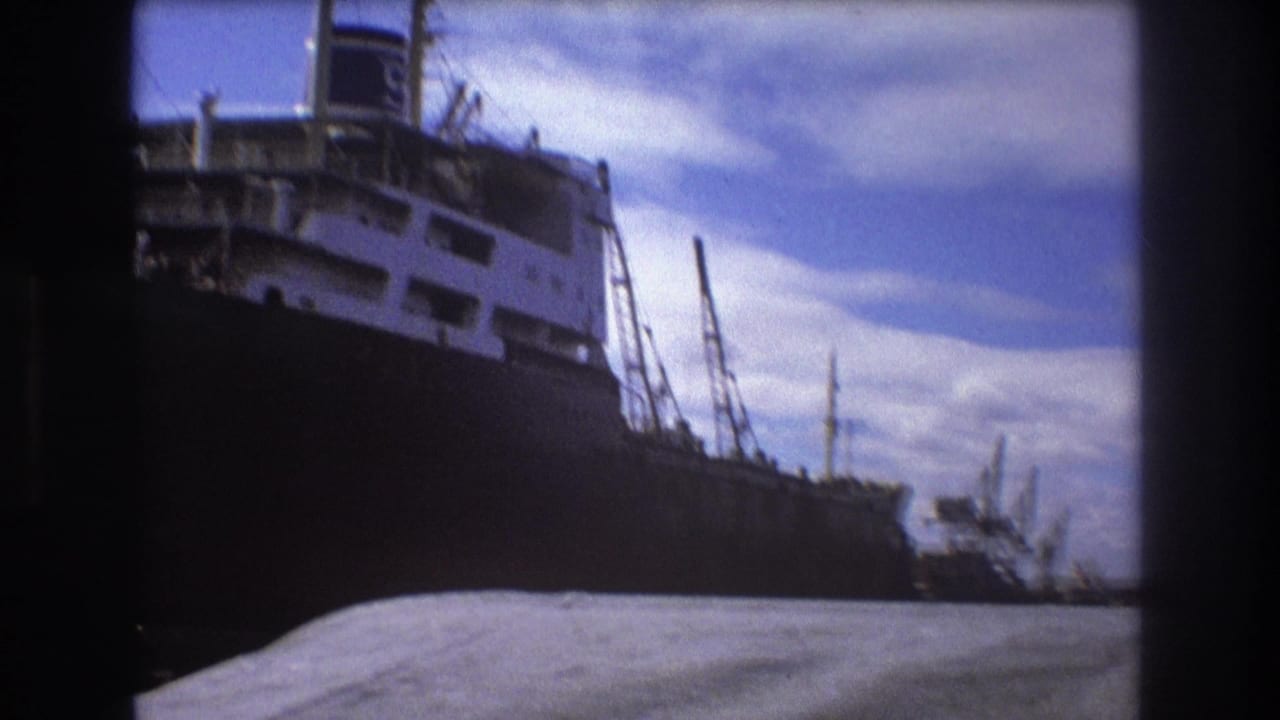 1975 Boston harbor frame showing the side and superstructure of a large freighter, industrial cranes, a tugboat-like vessel, and water in the foreground.