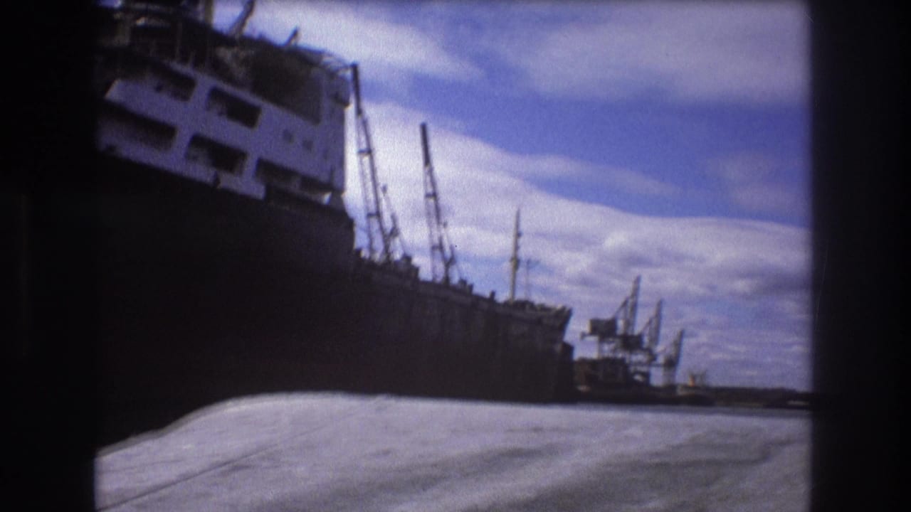 Color-shifted 8mm frame of a freighter bow in Boston harbor, with a small tugboat-like craft, cranes, water, and cloudy sky visible.