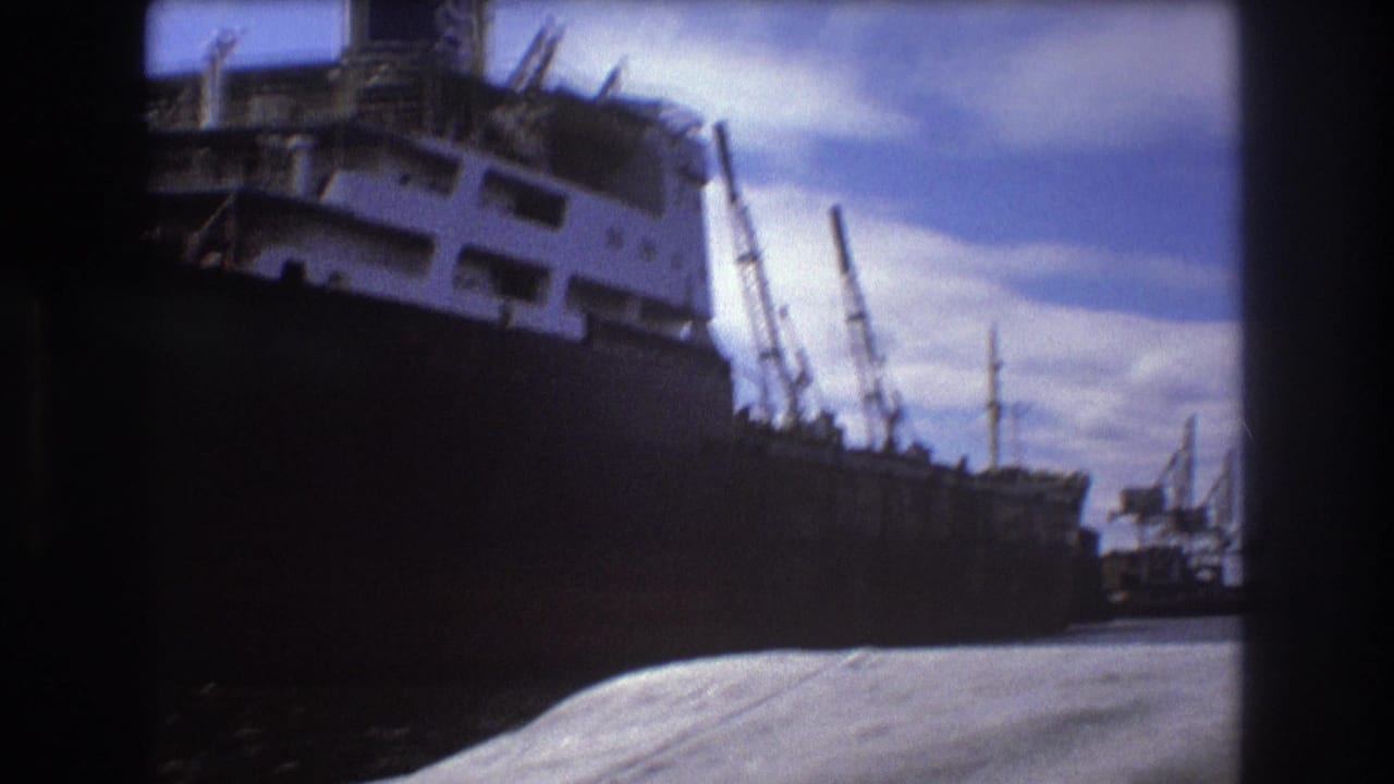 1975 Boston home-movie frame showing a large docked cargo ship with a dark hull, white superstructure, dock cranes, and bright sky.
