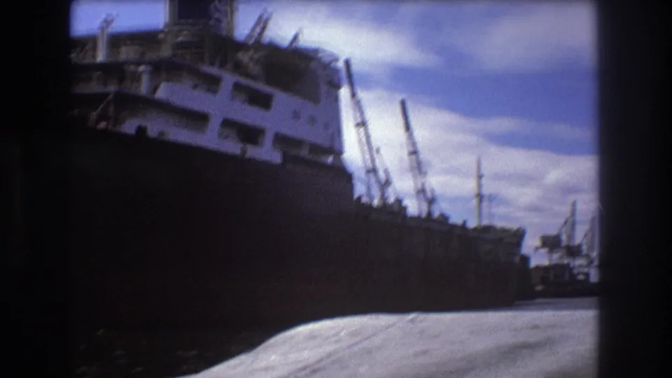 1975 Boston home-movie frame showing a large docked cargo ship with a dark hull, white superstructure, dock cranes, and bright sky.
