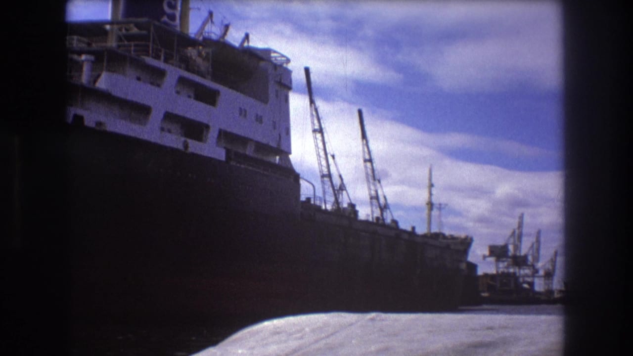 1975 Boston harbor view of a docked cargo vessel, its white upper decks, nearby cranes, and industrial waterfront under scattered clouds.