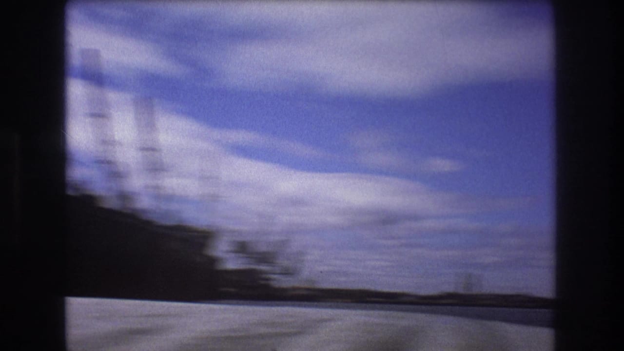 Handheld 1975 Boston harbor frame with motion blur across a dark ship form, bright foreground, distant shoreline, and clouded sky.