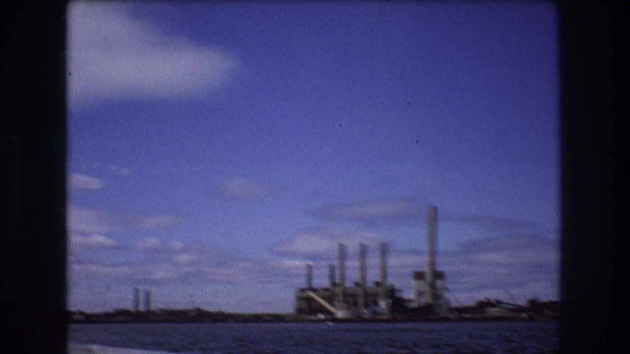 1975 Boston-area waterfront frame showing distant factory-like buildings and tall smokestacks across the water beneath a blue, lightly clouded sky.
