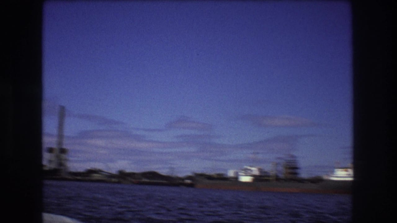 Soft 1975 Boston harbor 8mm frame showing water in the foreground, distant industrial shoreline, and a cargo vessel viewed from a moving boat.
