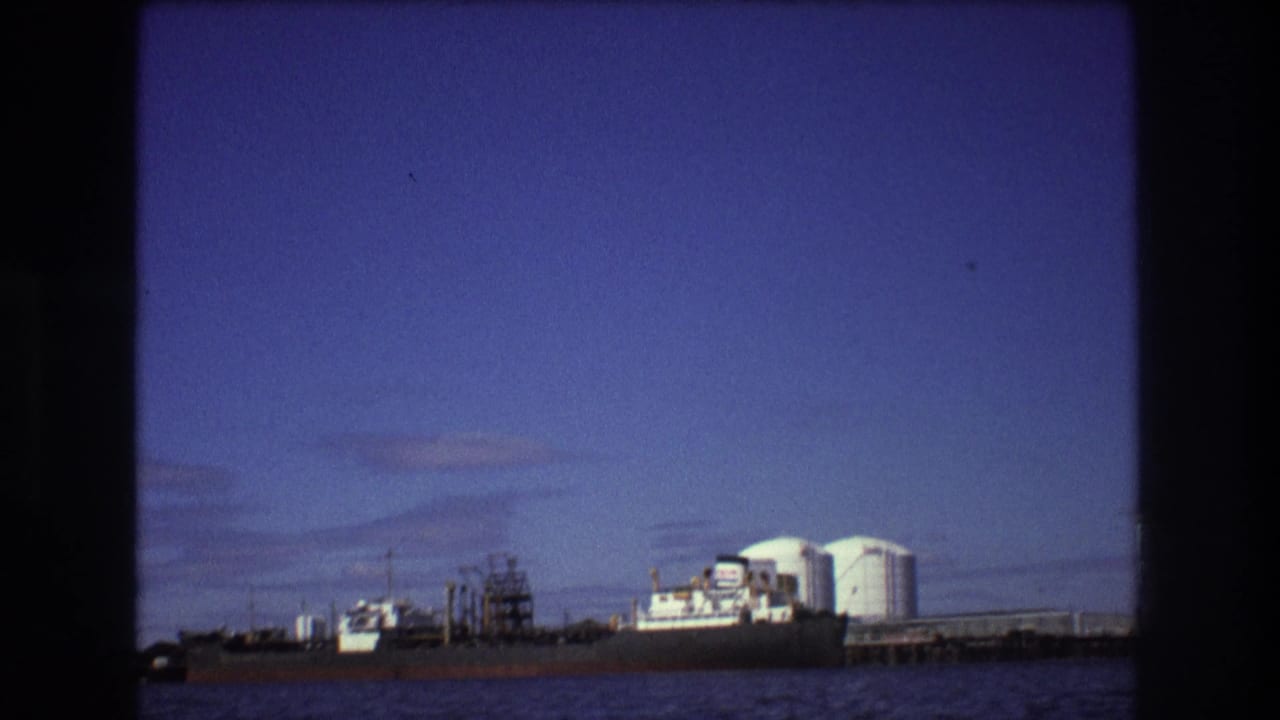 Cargo ship docked beside white storage tanks and pier structures across the water.