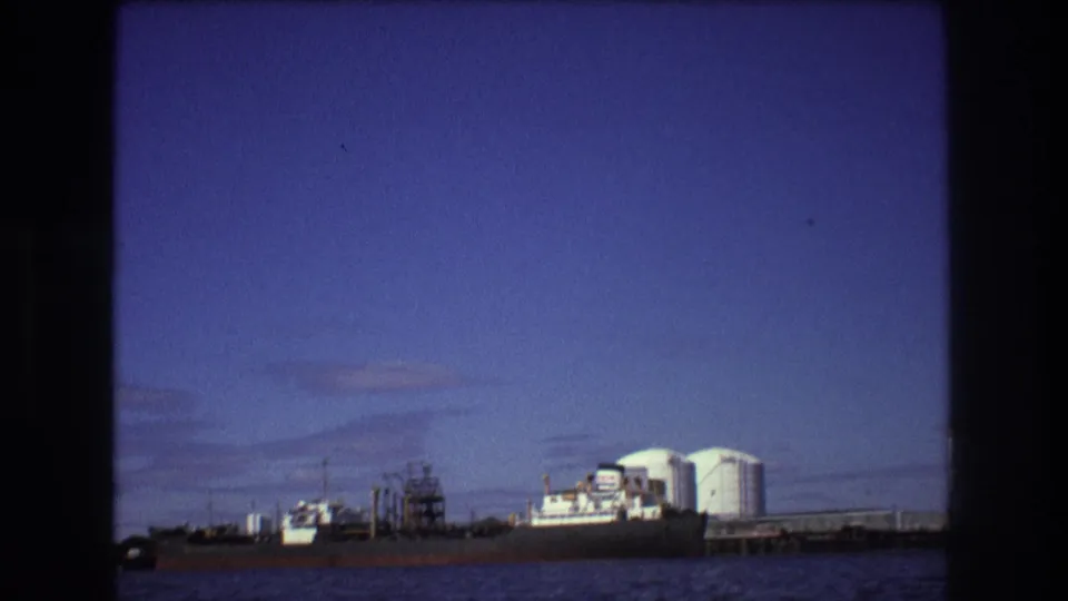 1975 Boston harbor frame with a docked cargo ship, white cylindrical tanks, and pier infrastructure seen across choppy water.
