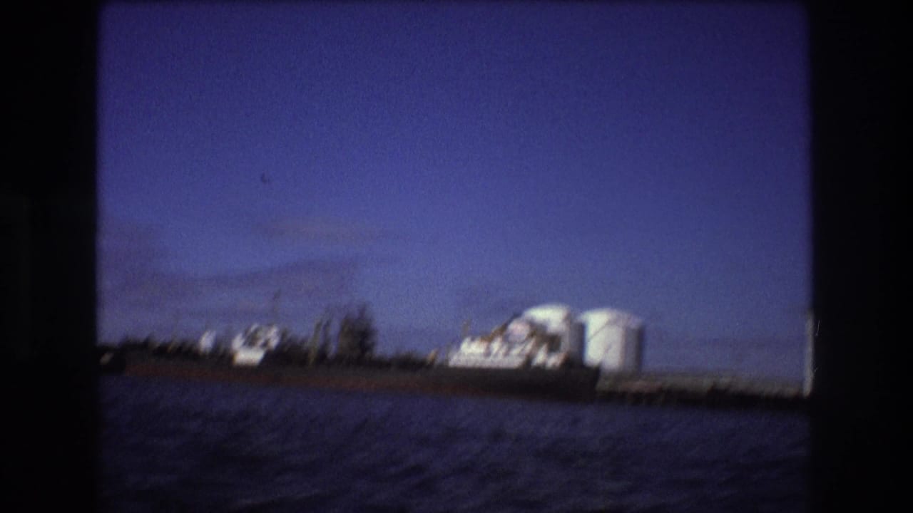 Wide 8mm view of Boston harbor in 1975 with an industrial ship at the pier, white tanks, and low waterfront structures across the water.