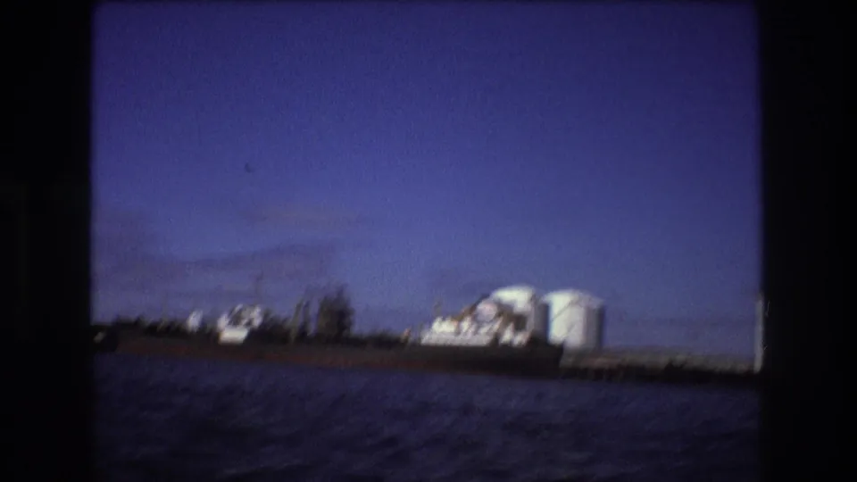 Wide 8mm view of Boston harbor in 1975 with an industrial ship at the pier, white tanks, and low waterfront structures across the water.