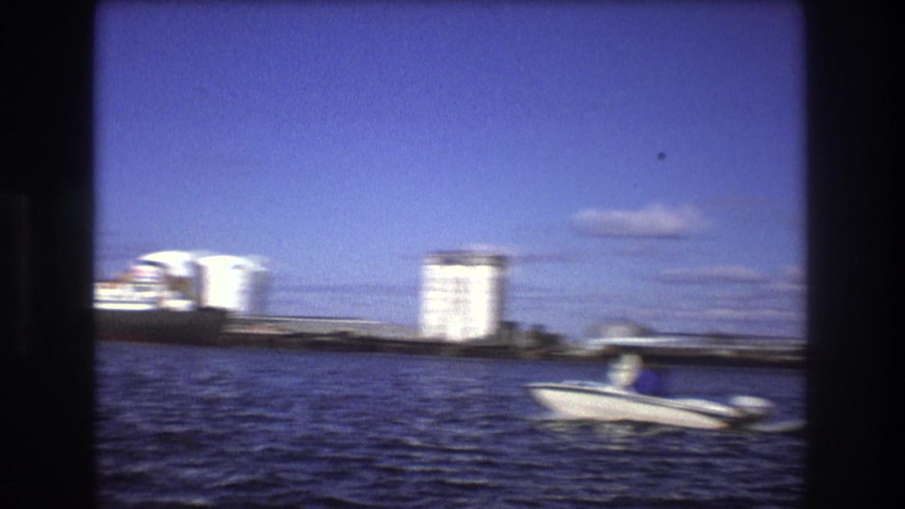 1975 Boston harbor home-movie frame showing a small motorboat with a visible passenger crossing in front of industrial tanks and waterfront buildings.