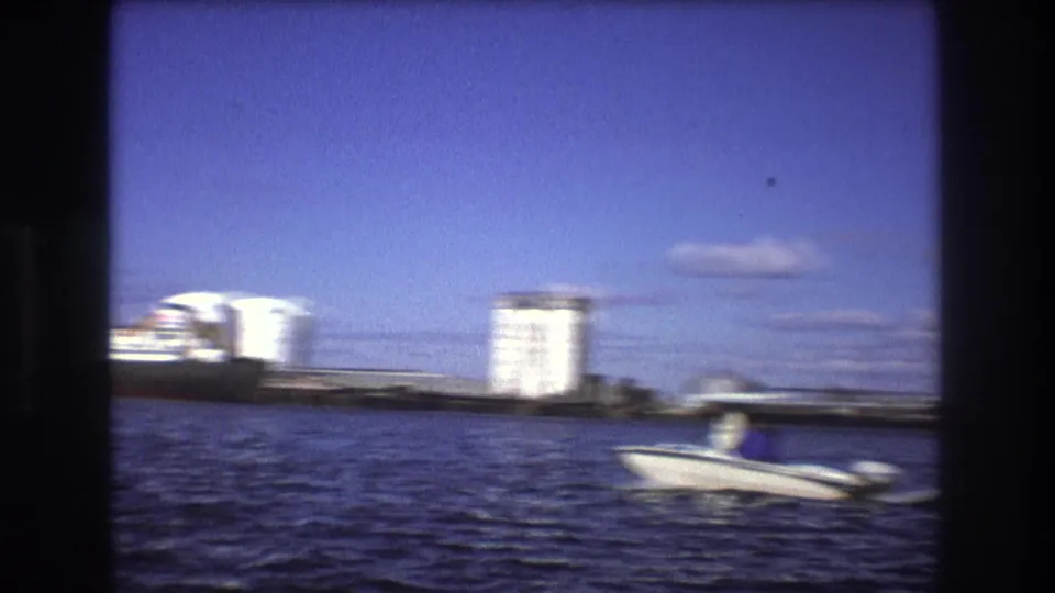 1975 Boston harbor home-movie frame showing a small motorboat with a visible passenger crossing in front of industrial tanks and waterfront buildings.