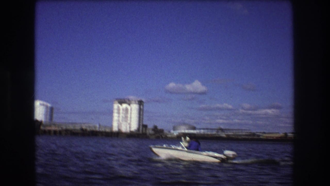 A small outboard motorboat crosses Boston harbor water in 1975, with tall storage silos and low industrial waterfront buildings under a blue sky.