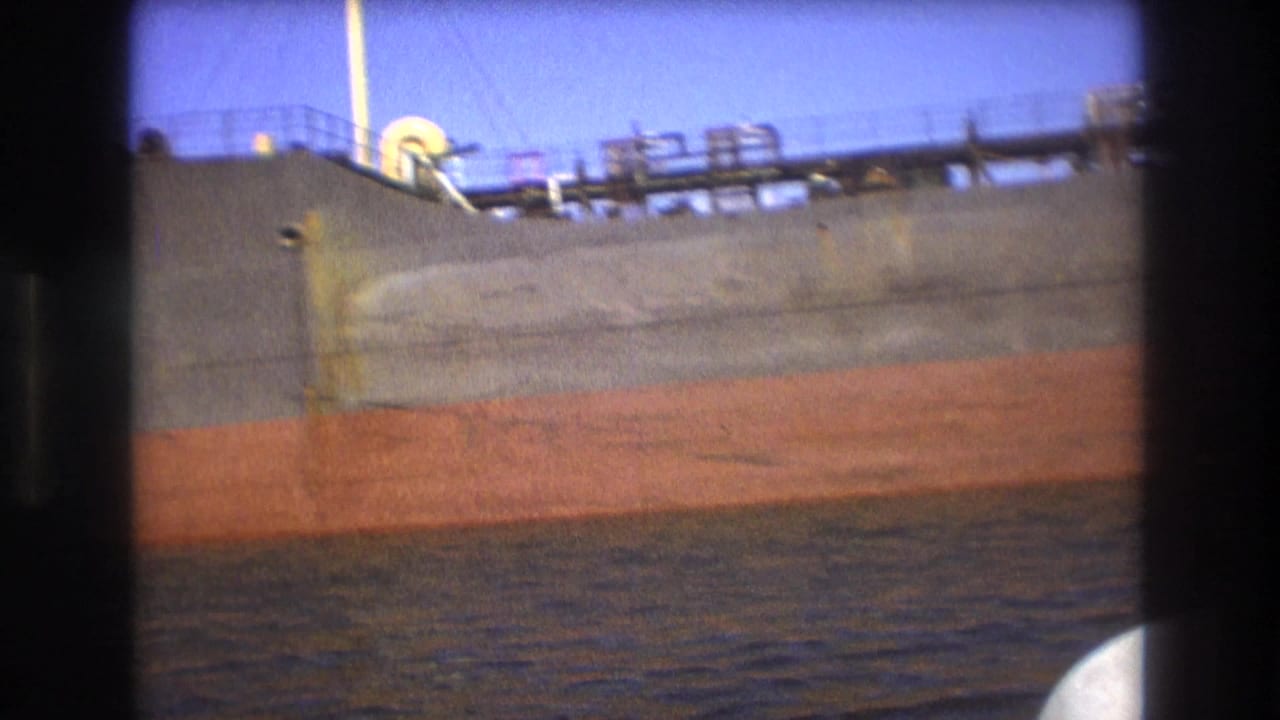Close view of a large freighter hull with a red lower band and deck equipment above the water.