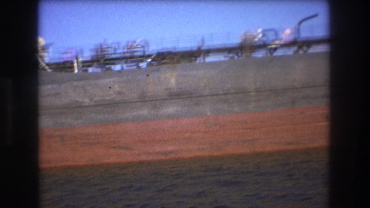 The camera continues past the freighter's side in Boston harbor, emphasizing the long gray hull, reddish lower section, deck fixtures, and rippling water.