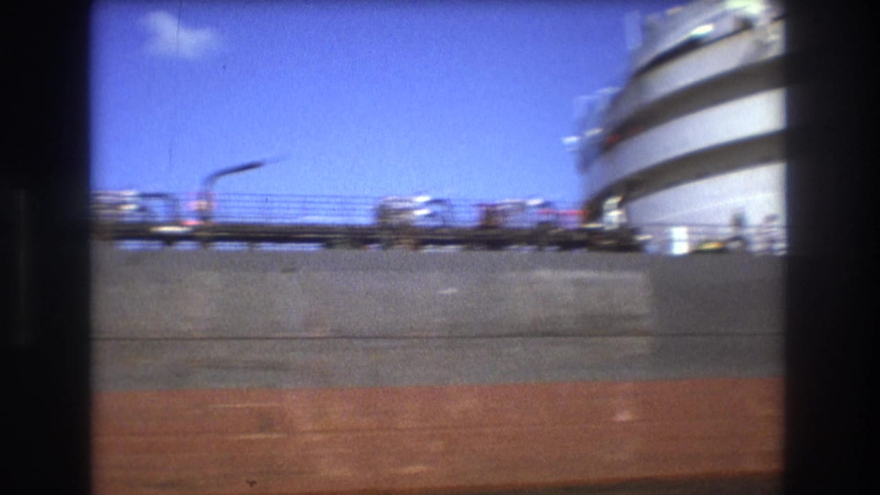A close harbor view shows the freighter's gray and red hull with deck railings, while a rounded dockside or vessel structure blurs along the right edge.