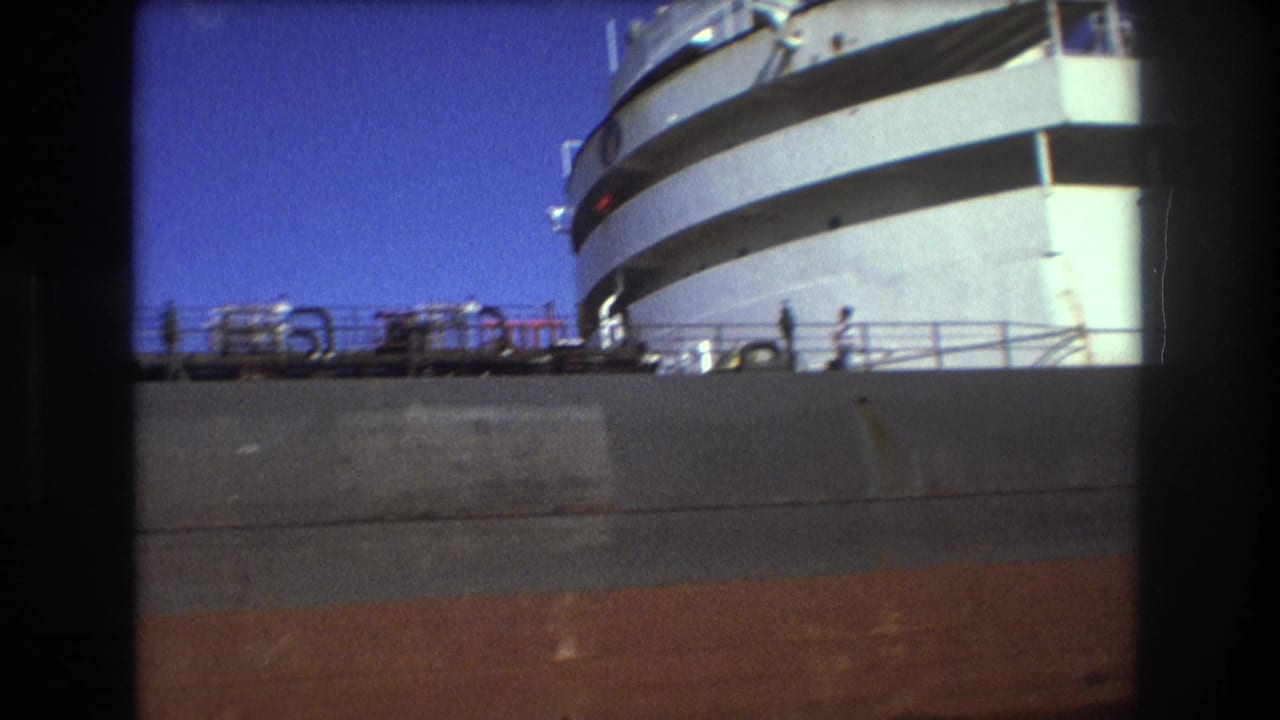 1975 Boston harbor frame with a large white passenger ship, dark bridge-window band, railings, and distant people seen across a dockside barrier.