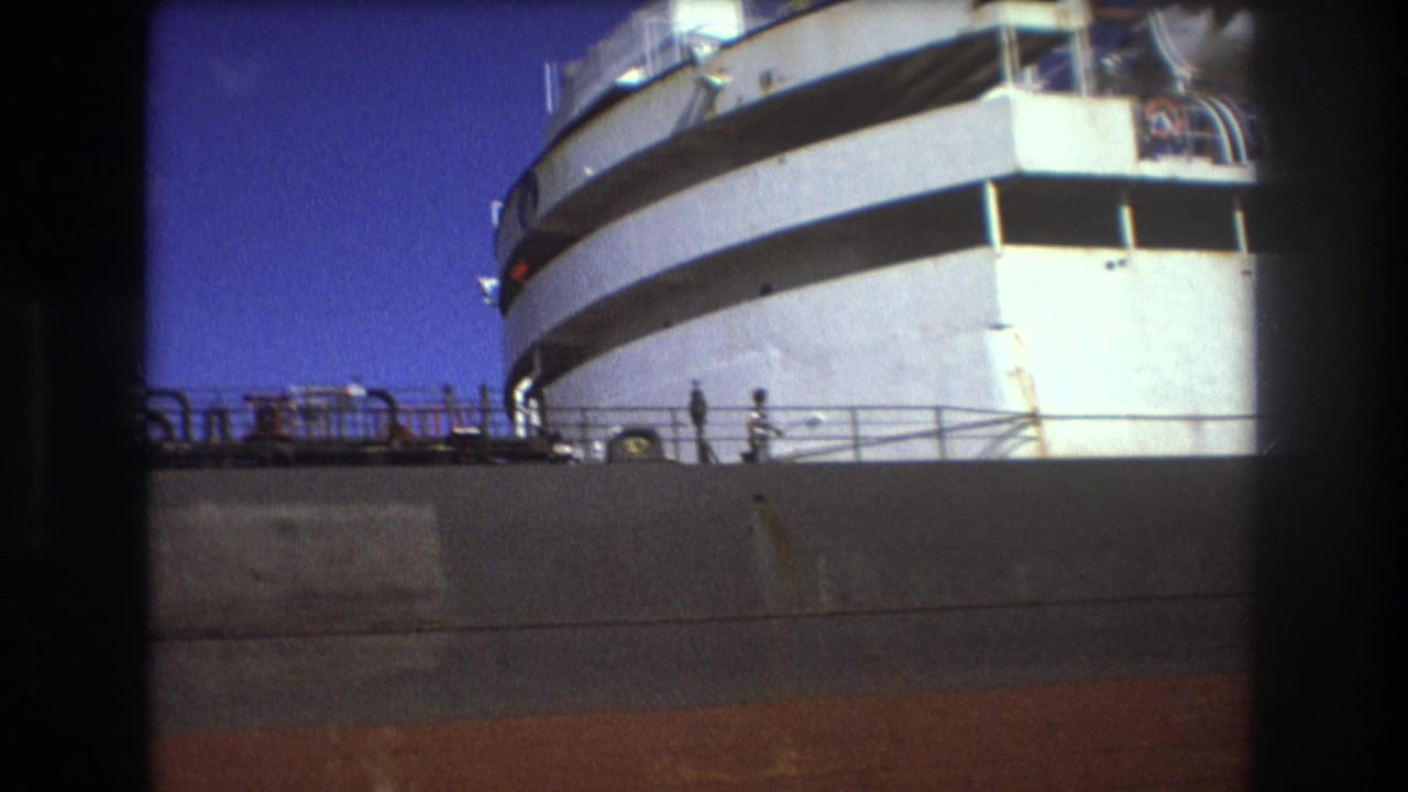 Low-angle 8mm view of a large passenger ship in Boston in 1975, with the white bow, upper decks, railing line, and tiny figures along the dock or deck area.