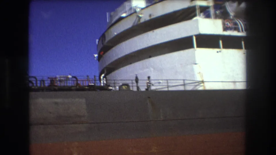 Low-angle 8mm view of a large passenger ship in Boston in 1975, with the white bow, upper decks, railing line, and tiny figures along the dock or deck area.