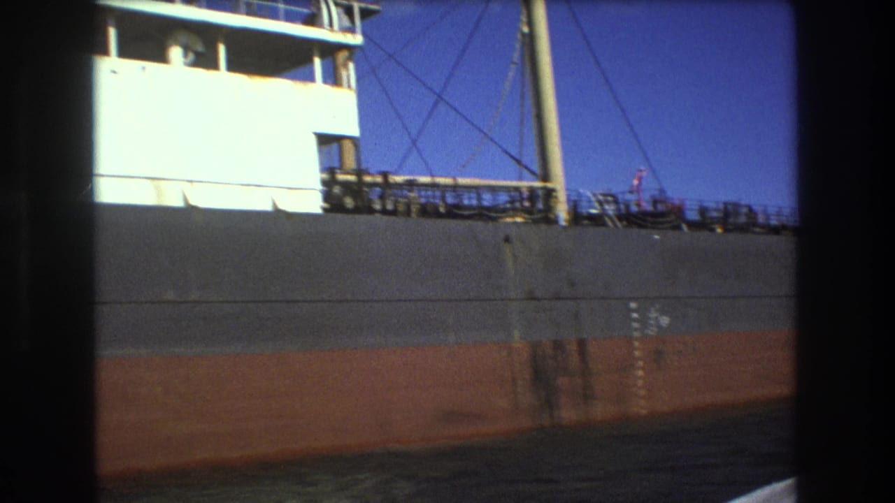 1975 Boston home-movie frame showing a large freighter passing close by, with a gray upper hull, rust-colored lower hull, deck railings, and the white superstructure partly visible.