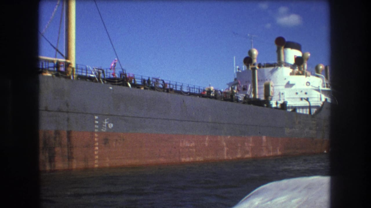 1975 Boston home-movie frame showing the midsection and stern-side superstructure of a large freighter, with deck details, smokestacks, and the filming boat edge in the foreground.