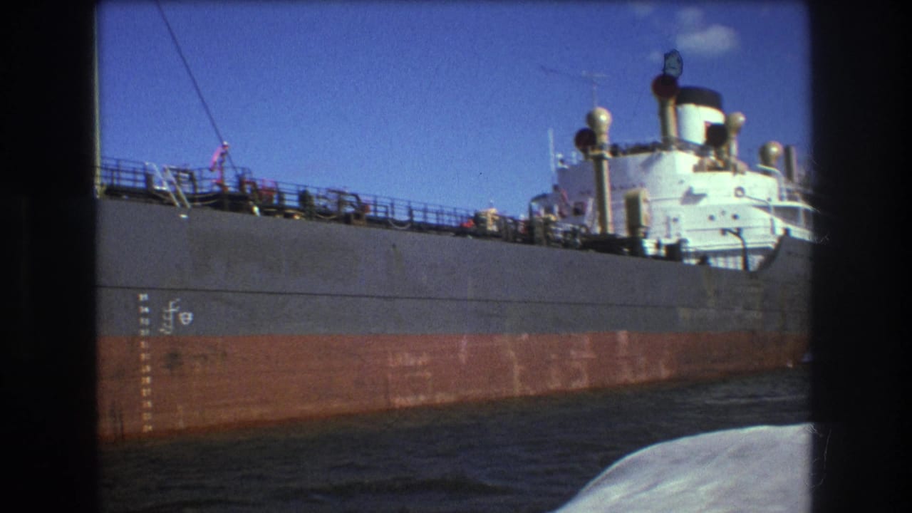 1975 Boston home-movie frame showing the freighter’s side, white bridge structure, railings, hull markings, and a rounded edge of the filming boat near the lower right.