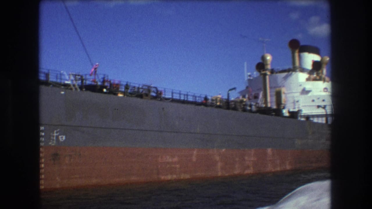 1975 Boston home-movie frame showing the freighter continuing past the filming boat, with the gray and rust hull, deck railings, white superstructure, and small distant deck figures visible.