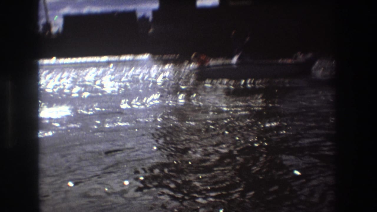 A small motorboat appears on shimmering water near dark waterfront silhouettes in home-movie footage identified as Boston, United States, 1975.