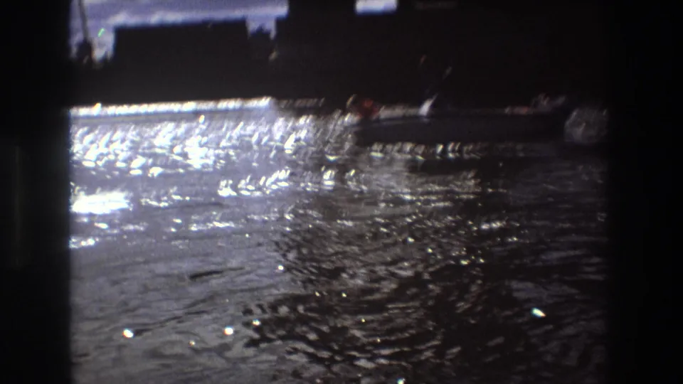 A small motorboat appears on shimmering water near dark waterfront silhouettes in home-movie footage identified as Boston, United States, 1975.