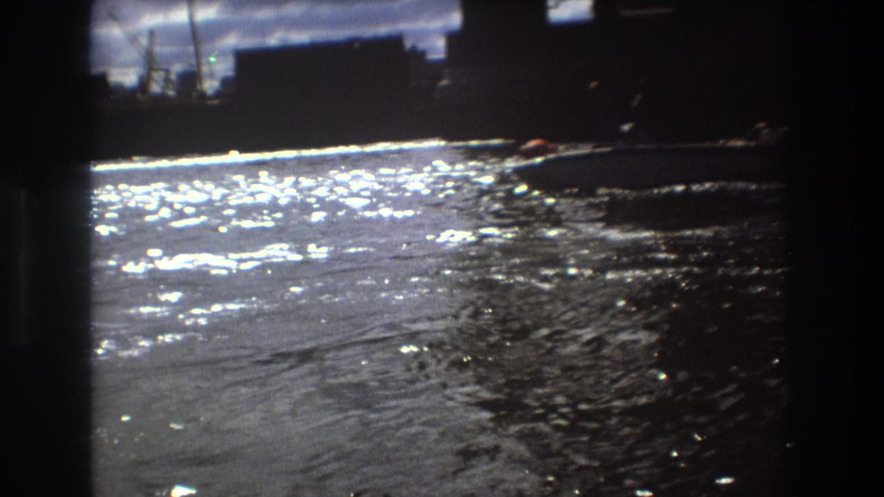 The frame shows rippling sunlit water with a small boat near the far right edge and dark waterfront forms in 1975 Boston home-movie footage.