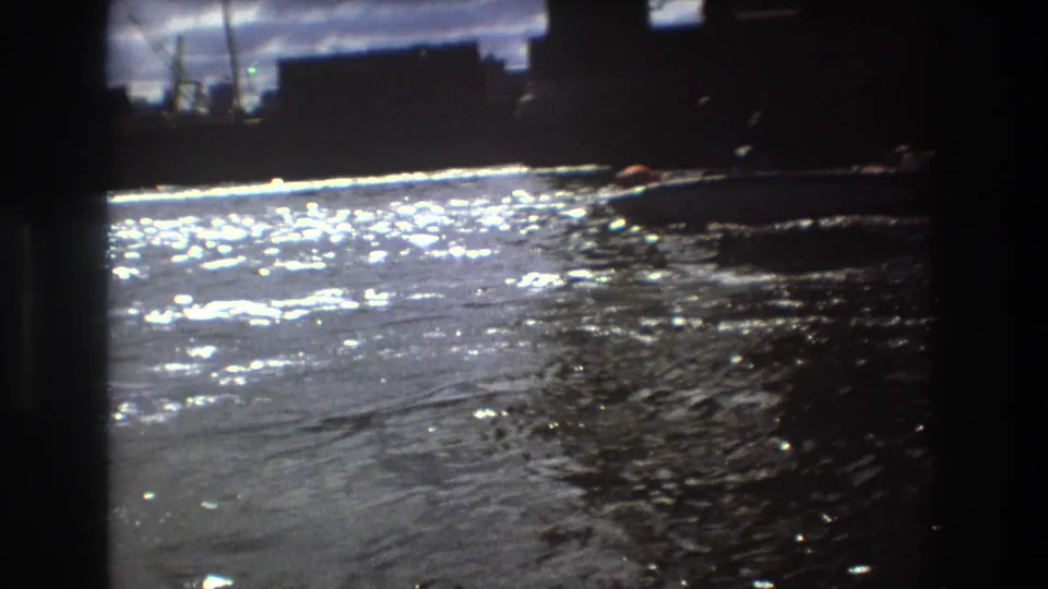 The frame shows rippling sunlit water with a small boat near the far right edge and dark waterfront forms in 1975 Boston home-movie footage.