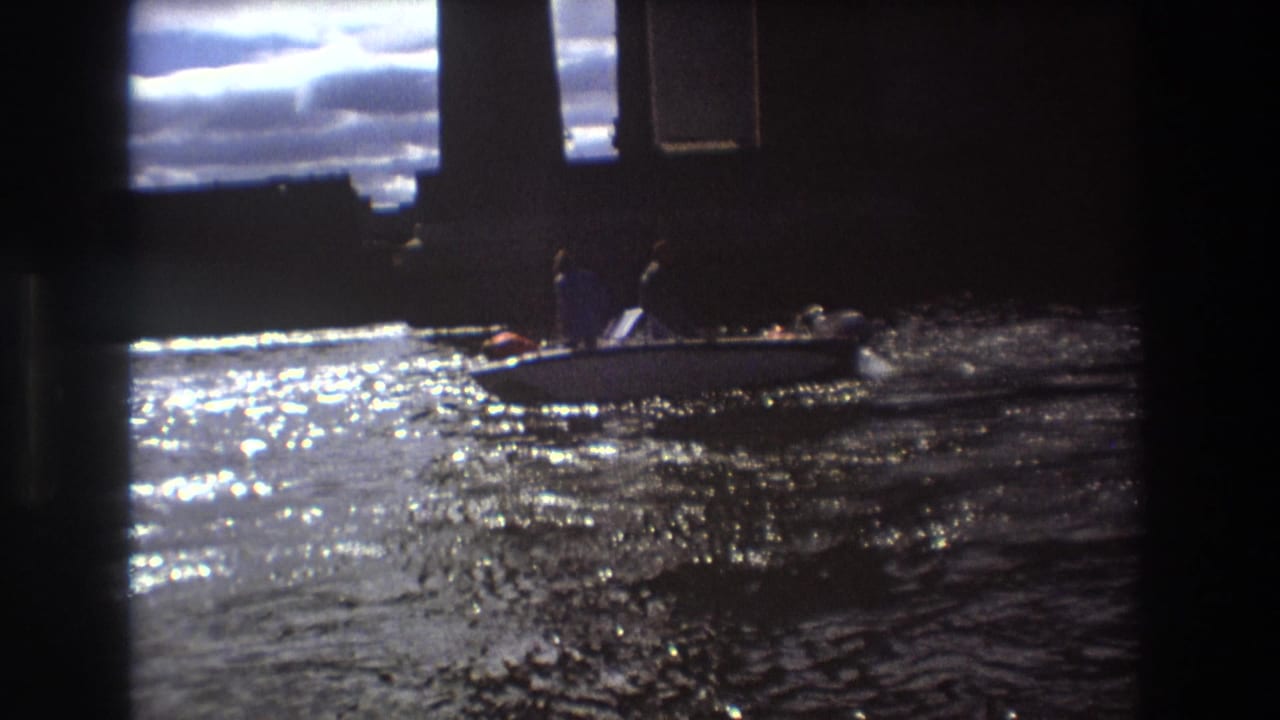 A small motorboat carrying distant passengers crosses sparkling water beneath dark vertical waterfront structures in archival 8mm footage from Boston metadata dated 1975.