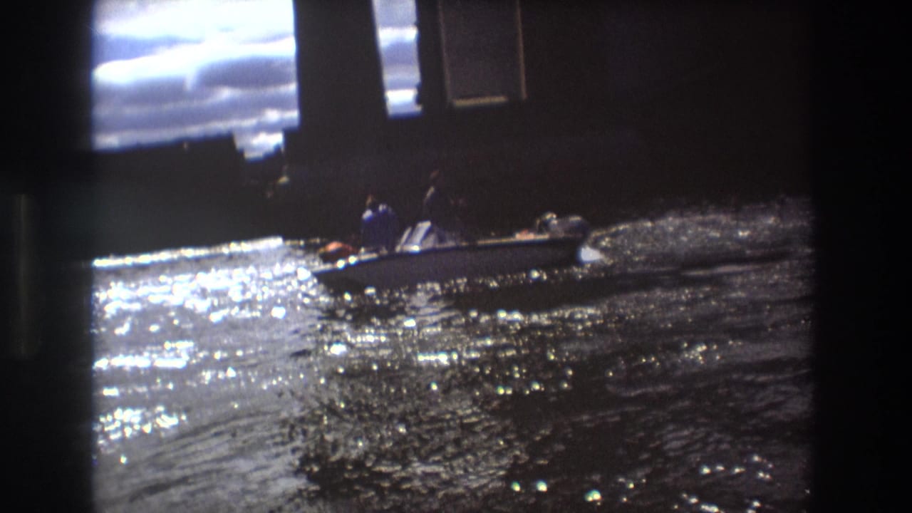 1975 Boston 8mm frame showing a small motorboat with people aboard moving across glittering river water near large bridge supports.