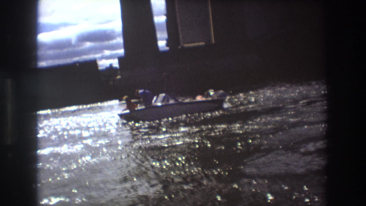 1975 Boston home-movie frame with a small open boat passing across bright, rippled river water beside shadowed bridge structure.