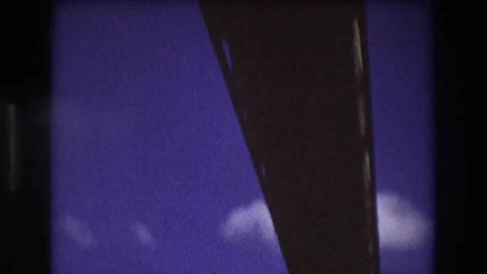 Soft 8mm frame from the 1975 Boston clip showing a dark bridge or boat structure cutting across blue sky with small clouds visible below.