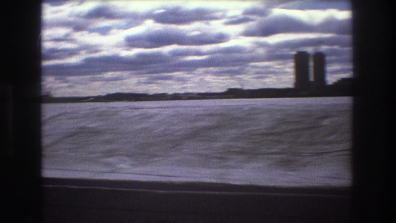 1975 Boston boat-view frame showing rippled water, a cloudy sky, low shoreline, and two tall buildings near the right side of the horizon.