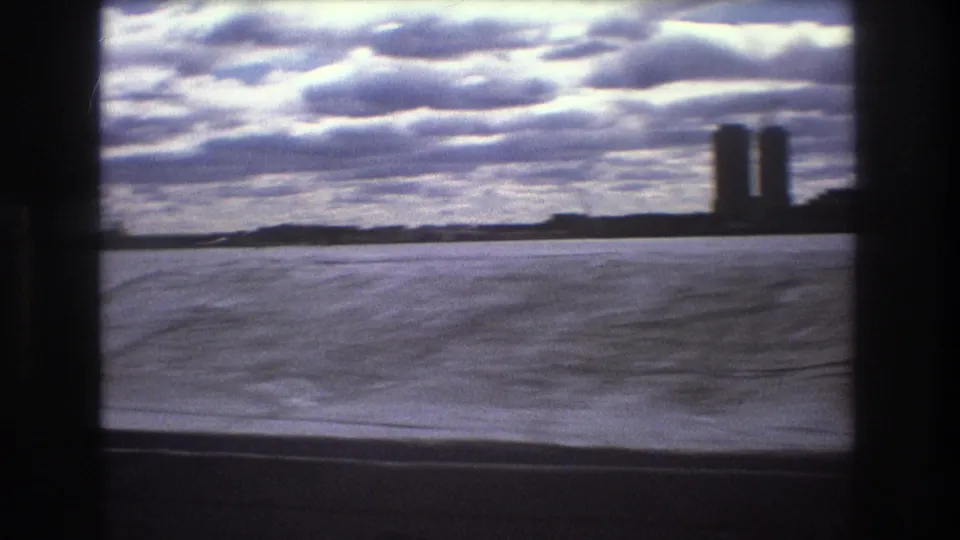 1975 Boston boat-view frame showing rippled water, a cloudy sky, low shoreline, and two tall buildings near the right side of the horizon.