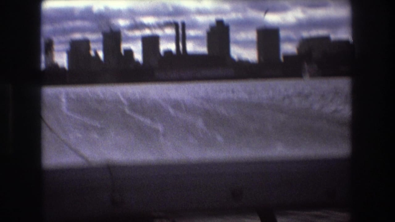Boston, 1975, with a dark waterfront skyline, tall buildings, possible industrial stacks, and rippled bay water viewed through shadowed boat framing.